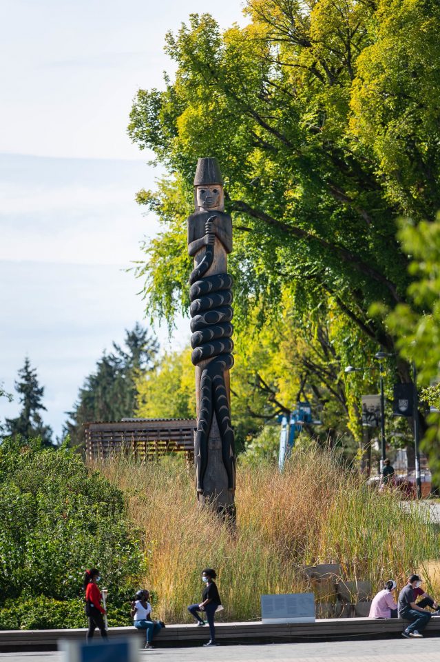 sʔi:ɬqəy̓ qeqən (double-headed serpent post), Brent Sparrow, Musqueam artist's work set in a treed area with some students milling around casually